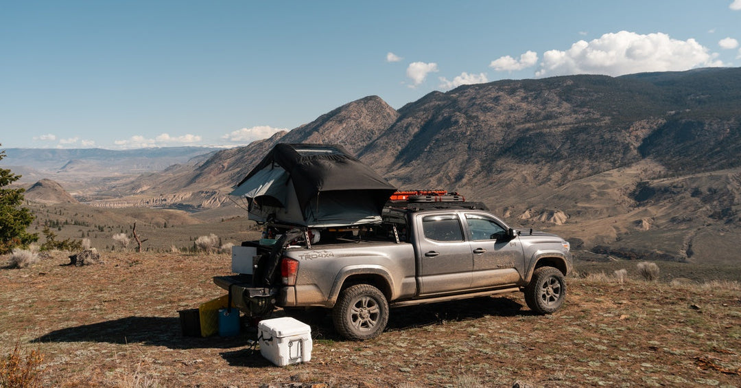 A pickup truck with a camping tent on top and multiple accessories alongside it parked on a flat surface next to a mountain.