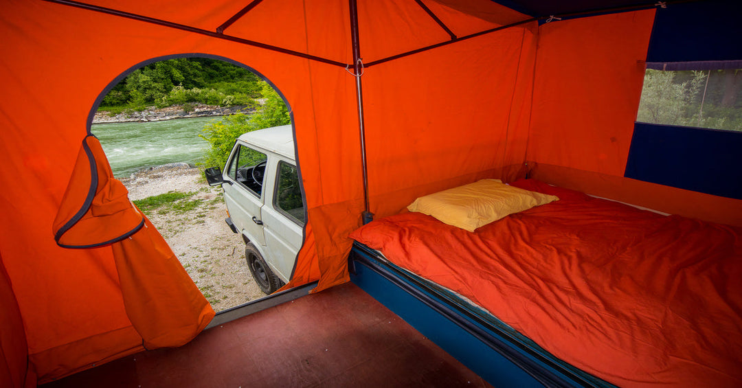 Orange camping tent on top of the back of a truck with a bed inside and a view of a river with greenery around.