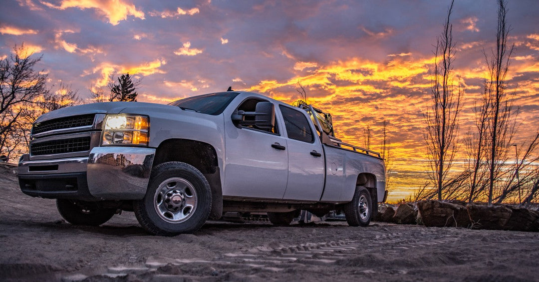 Large white pickup truck with the headlights on, parked on a dirt road with a beautiful sunset in the background.