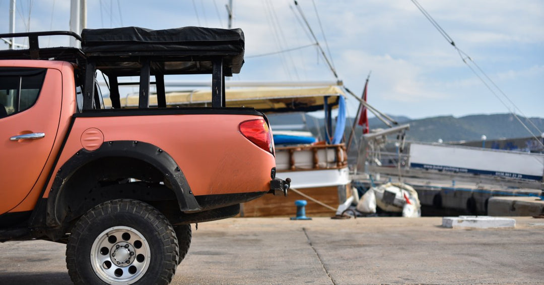 Bed of an orange pickup truck with a black back rack carrying a black cover, parked on a sea pier next to a boat.