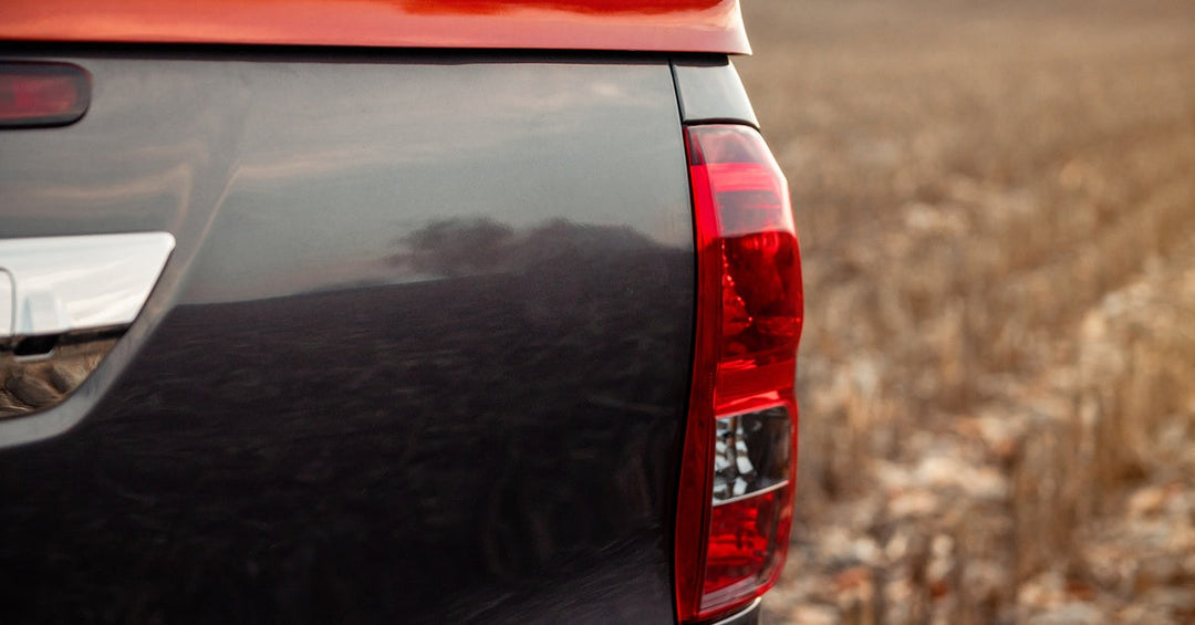 Close-up to the stop light on the back end of a dark grey pickup truck with a hard red cover parked in a field.