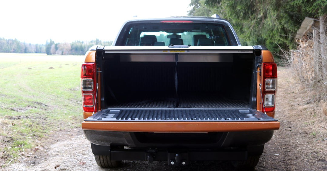 Orange pickup truck with a bed cover and an open tailgate parked on a dirt road next to a large space covered in grass.