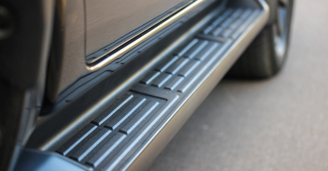 Close-up of a black running board with an antislip detail attached to the side of a black vehicle parked on concrete.
