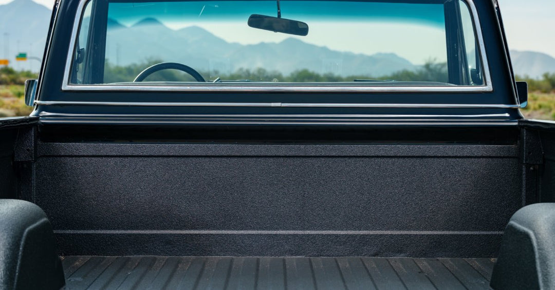 A close-up of the bed of a vintage pickup truck with a black plastic finish parked outdoors with a mountain view.