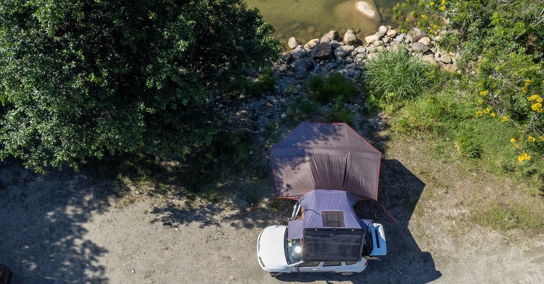 An aerial view of a white vehicle with a large awning on top and a tent, parked on a dirt road next to a body of water.