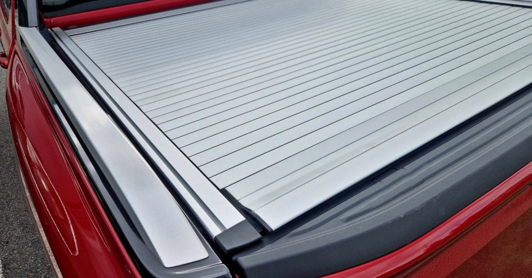 A close-up of an aluminum bed cover with a black trim on the bed of a red pickup truck parked next to a car.