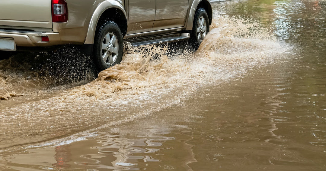 A brown pickup truck with two sitting cabins, driving through a flooded street and creating small waves.