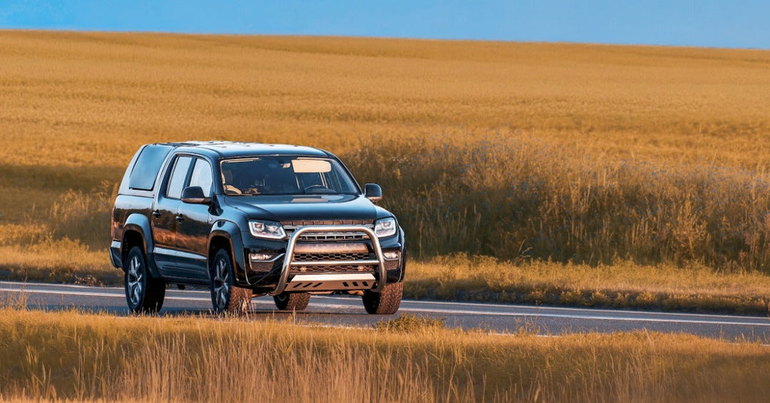 A pickup truck with a metal front bumper and a bed truck, driving on a concrete road next to tall, dry grass.