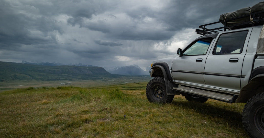 A grey pickup truck with a black metal roof rack carrying a black bag, parked in a green landscape on a cloudy day.