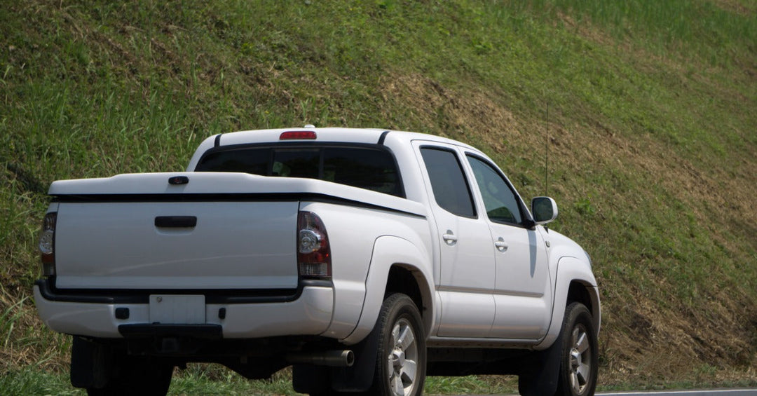 A white pickup truck with a bed cover driving on a concrete road next to a hill covered in grass on a sunny day.