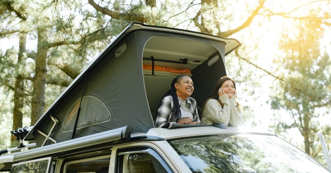 Two women looking up inside a grey foldable tent on the roof of a white truck parked next to multiple trees.
