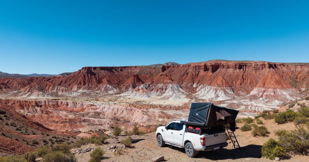 A white pickup truck with a bed tent and a small ladder, parked on the side of a large canyon on a sunny day.