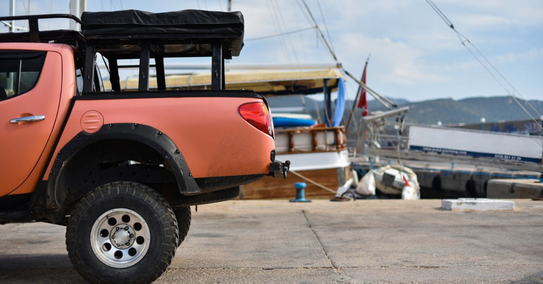 The back of an orange pickup truck with a black rack holding a large bag parked next to a pier with floating boats.
