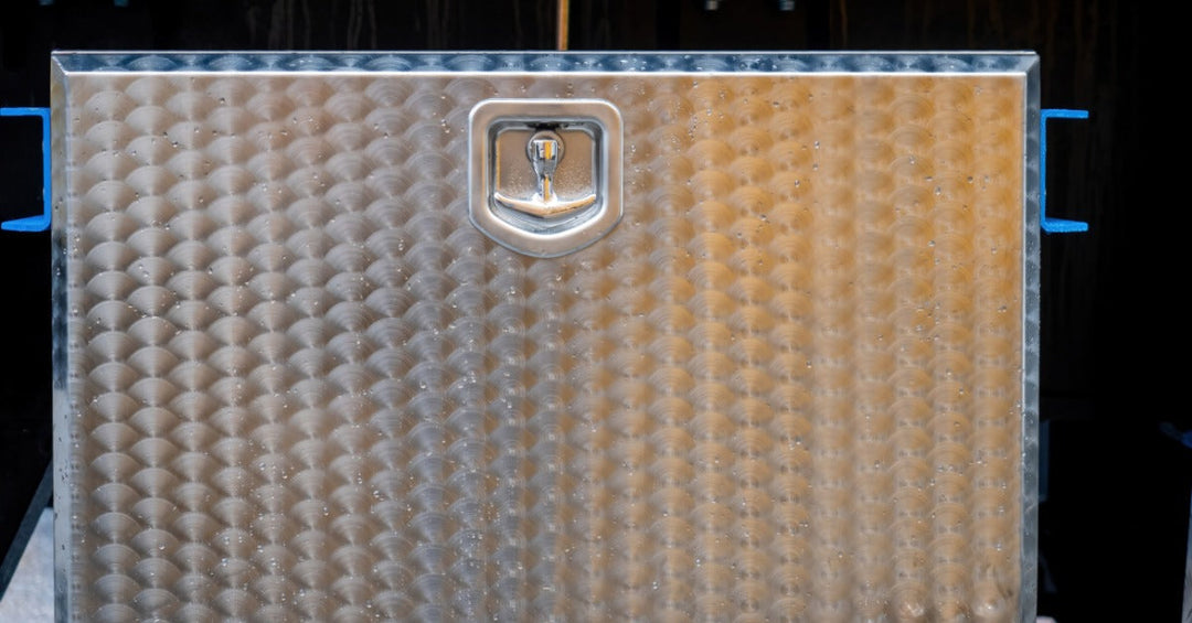 A metal toolbox with a patterned design and a small handle on top placed on a flat surface with bolts in the background.
