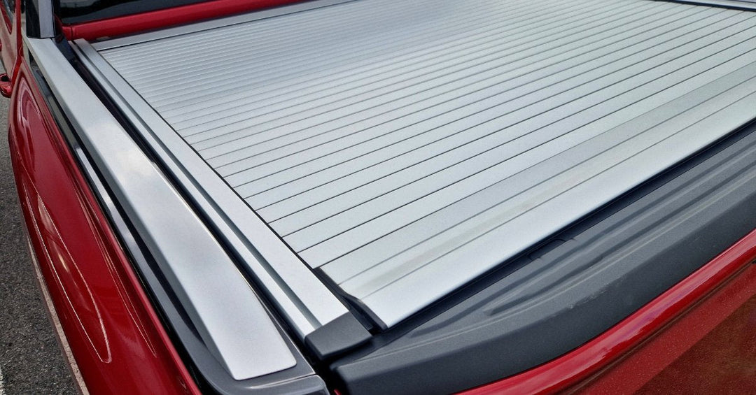 A close-up of the back of a red pickup truck with a black, foldable Tonneau cover parked in a parking lot.