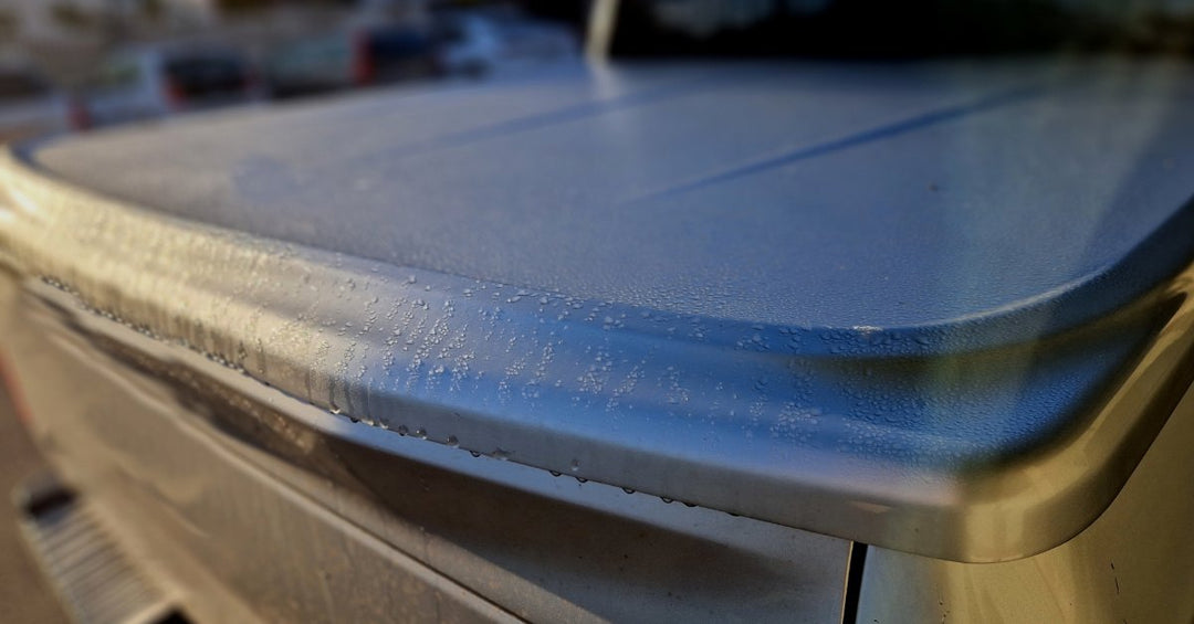 A close-up of a soft cover over the bed of a grey pickup truck covered in small water drops parked outdoors.