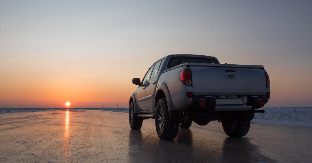 The back of a white pickup truck with a bed cover and silver wheels, driving towards a sunset on a wet pavement road.
