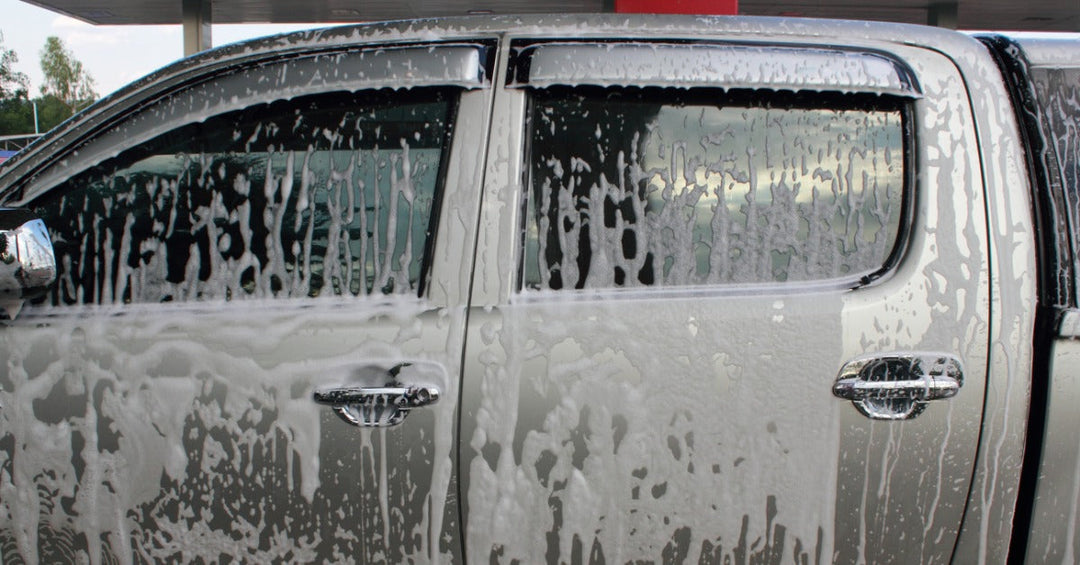 A close-up of the side of a grey pickup truck with a bed cap and tinted windows covered in white soap foam.