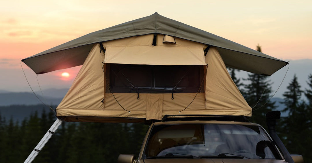 A large roof-top tent on top of a bronze pickup truck parked on a mountain with trees and a sunset in the background.