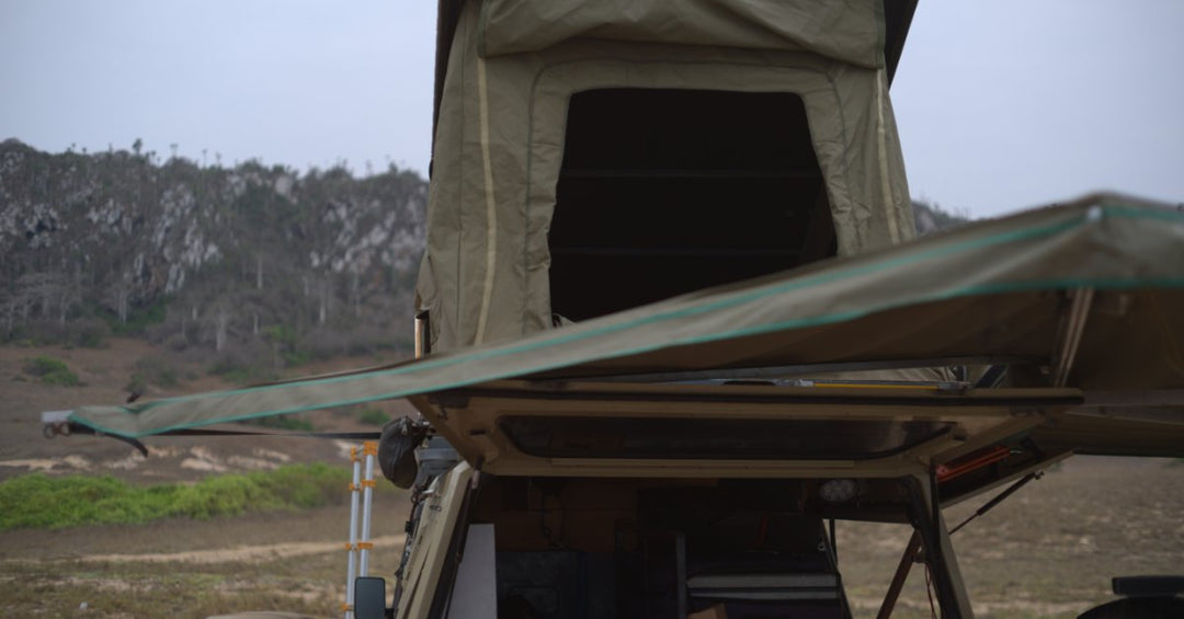 A close-up of a green awning next to a tent on top of a pickup truck parked on a cloudy, deserted landscape.