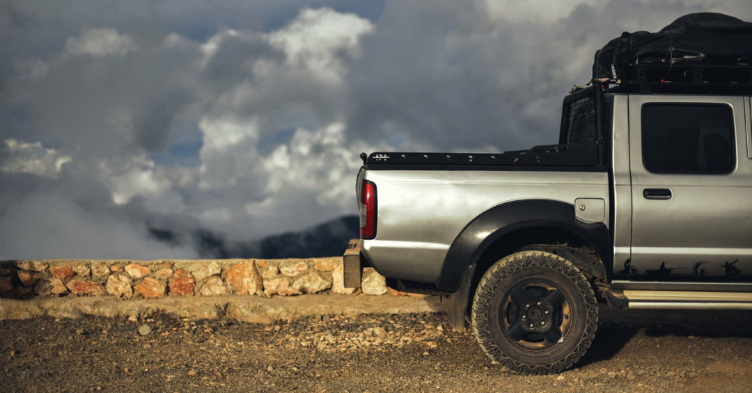 A side view of the back of a grey pickup truck with a metal rack in the bed parked next to a small rock wall.